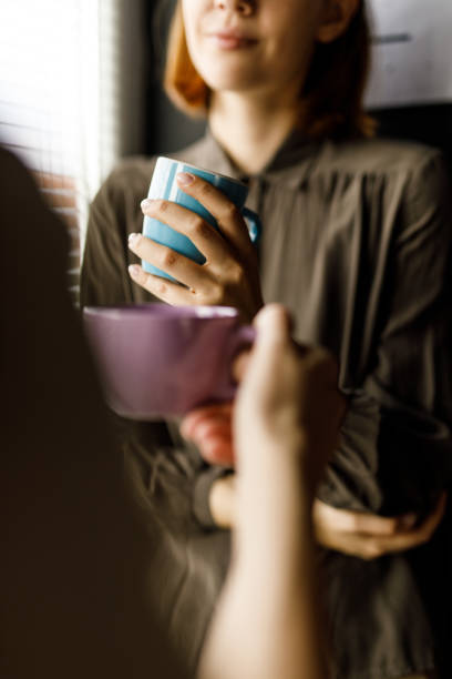 Two young women standing by the window, having cups of coffee and chatting