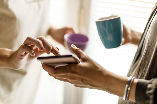 Close up shot of two unrecognizable women standing by the window, having coffee and looking at something online via smart phone.