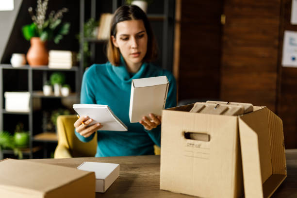 istock Young businesswoman sitting at her desk and checking her notes while sorting packages and boxes 1455776041