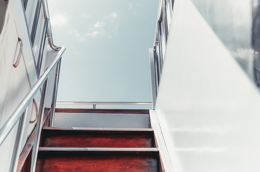 A shot captured from the lower deck when climbing a wooden stairway and shiny metallic railings leading to the upper deck of a white sailing yacht; on a warm sunny clear sky day in the Maldives island