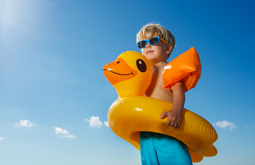 Happy little blond boy with inflatable yellow buoy, shoulder straps wear sunglasses close-up from low angle