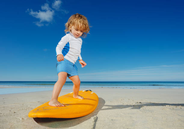 Little blond toddler girl already practicing and posing balancing standing on the surfboard on the beach