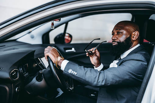 A view of a sophisticated black male, in selective focus, with one hand on the steering wheel and the other on the sunglasses he is going to put on, he's wearing an elegant tailored grey-scale suit