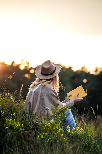 Caucasian woman wearing poncho and cowboy hat in nature. Relaxation with book during sunset