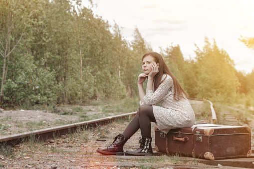 Teen girl in stylish beige dress sitting on vintage suitcase on abandoned railway in spring forest, lonely, looking away. Fashionable lady in retro image. Travel vacation concept. Copy text space