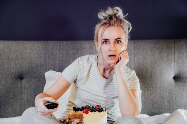 istock Emotionally stressed, shocked woman eating cake and holding a remote control while watching TV in bed. Scary horror movie or terrible news, scare in her eyes Overeating, Jamming of negative emotions. 1455778907