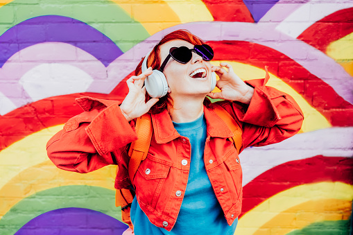 Emotional stylish woman in bright clothes wearing wireless headphones, listening to music and singing on rainbow graffity on the brick wall background. Fashionable hipster lifestyle.