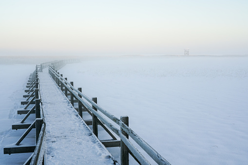 Snowy wooden boardwalk on frozen lake to birdwatching tower at sunset, light fog landscape