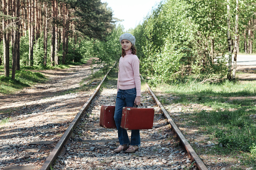 Serious teen girl in stylish french image standing with old suitcases on railway in spring forest, looking at camera. Traveler young lady in retro image waiting. Travel vacation concept. Copy space