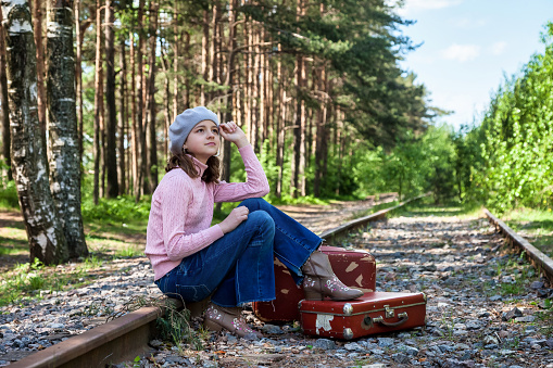 Smiling teen girl in stylish french image sitting on old suitcase on abandoned railway in spring forest, looking up away. Traveler young lady in retro image. Travel vacation concept. Copy text space