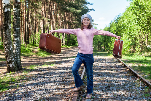 Happy smiling teen girl in stylish french image standing with old suitcases on railway in summer forest, looking at camera. Traveler young lady in retro image. Travel vacation concept. Copy space