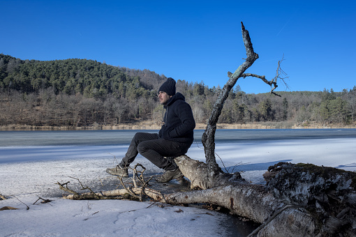 Middle-aged male hiker admires the landscape while sitting on a fallen tree in the frozen lake on a sunny day