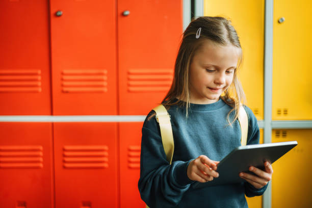 schoolgirl with digital tablet standing against locker in corridor stock photo