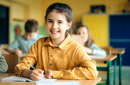 Side view of a schoolgirl sitting at classroom desk and writing in her workbook. Elementary schoolgirl studying in classroom.