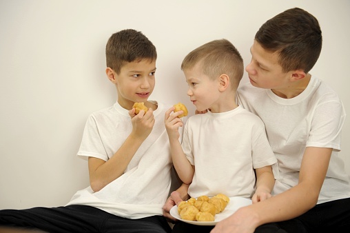 3 brothers in white t-shirts eat custard cakes sweets They look at each other smiling delicious dessert vacation family three boys sweets cakes home cooking white background