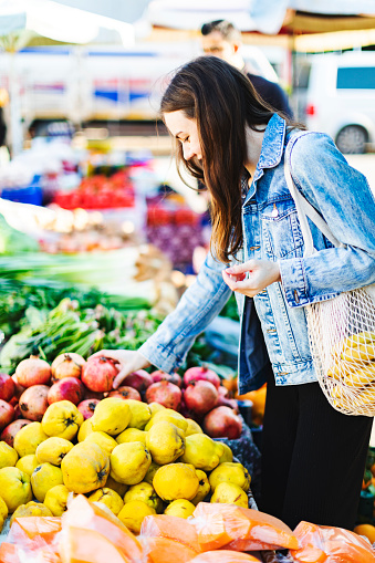 Young adult woman buying pomegranate at market with reusable bag