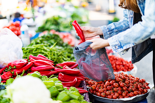 Young adult woman buying red bell pepper at market with reusable bag