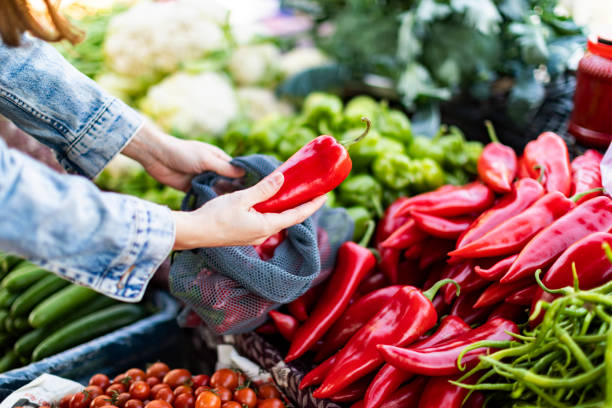 Young adult woman buying red bell pepper at market with reusable bag stock photo