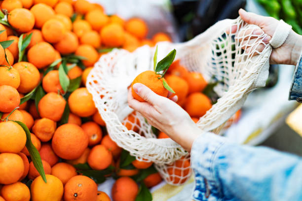 Giovane donna adulta che compra mandarino al mercato con borsa riutilizzabile - foto stock