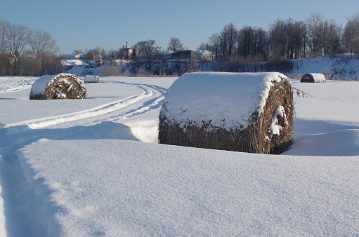 a rolled haystack under the snow against the background of the village and the blue sky