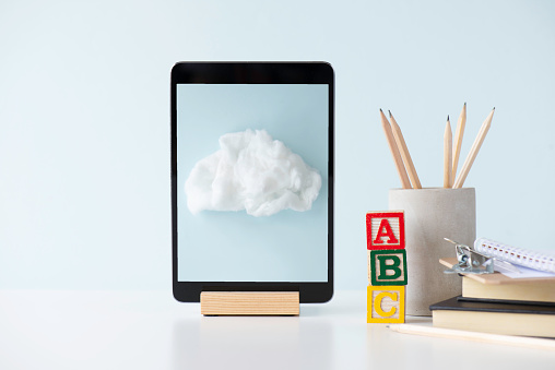 Front view of a white desk with a digital tablet, pen holder with pens, notepad,paperclip, and toy blocks with the letters A,B,C in front of light blue wall. There is an image of a cloud  on device screen. Representing e-learning.