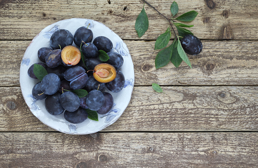 Fresh blue plums in bowl on wooden table with leaves and twig, halved plum with seed as decoration. Top view. Real photo taken from above.