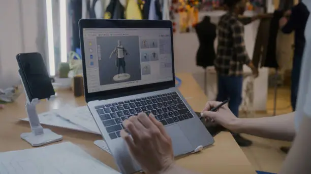 Female fashion designer develops future design of clothes in 3D modeling program on laptop using digital tablet and stylus. She works in atelier workshop. Fashion and technologies in business concept.