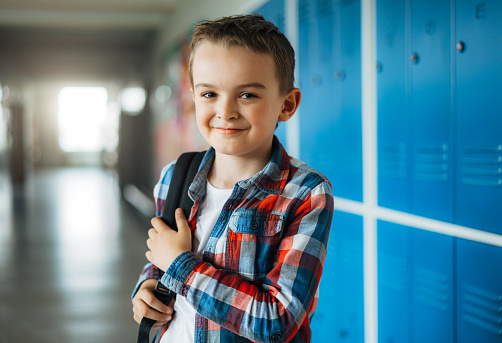 Portrait of a happy elementary schoolboy with schoolbag standing in school corridor in front of lockers. Smiling young boy looking to camera while standing in school.