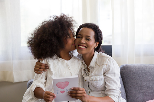 Child girl hugging and kissing her mother and holding happy mother day paper card at home