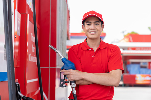 Asian attendant service male worker refuelling car at gas station. Smiling assistant man wear red uniform and red hat refuelling car at petrol station