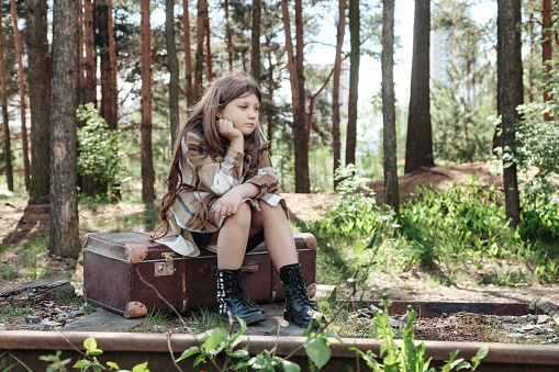 Pensive little girl in stylish coat sitting on vintage suitcase on abandoned railway track in spring forest, sad. Traveler child in retro image outdoors. Bad travel vacation concept. Copy text space