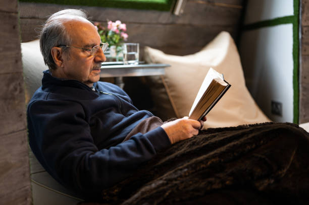 istock Elderly man with a mustache and reading glasses covered with a soft blanket sits on a bean bag and reads a book in cozy interior of wooden house 1455832965
