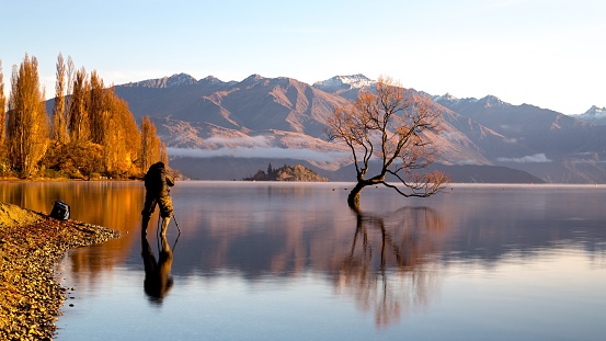 A scenic view of a person standing by the shore of a lake taking picture of the mountain in the back