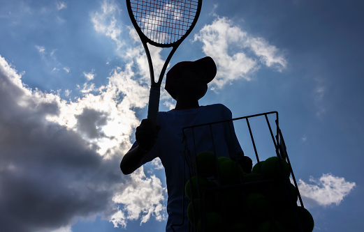 Tennis player near a basket of balls with his racquet against the light