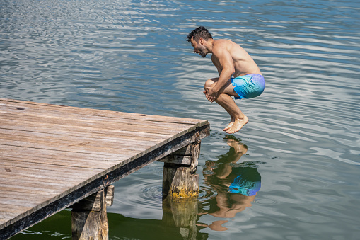Young Caucasian man jumping in the lake from wooden pier.