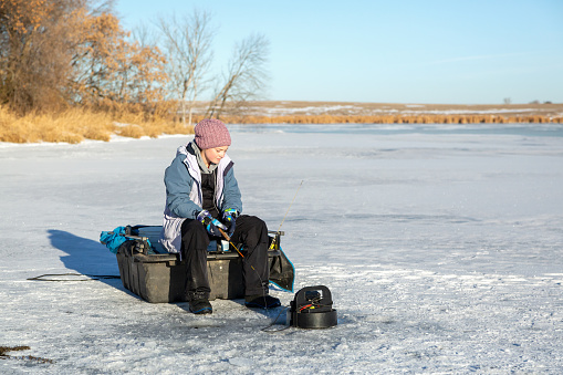 Girl sitting on ice fish house sled as she fishes on a frozen lake in winter.
