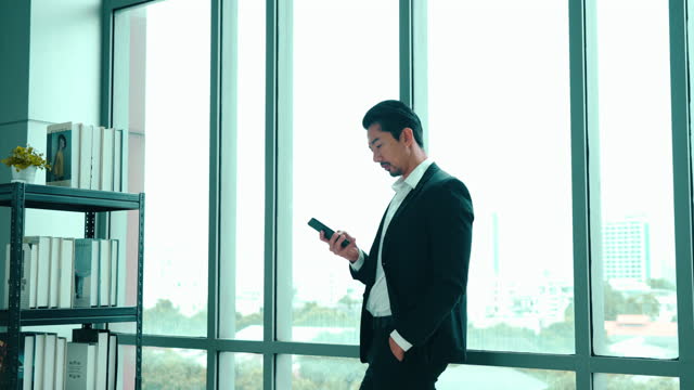 Asian young adult businessman is leaning against mirror wall and using smartphone to watch video online during rest time.