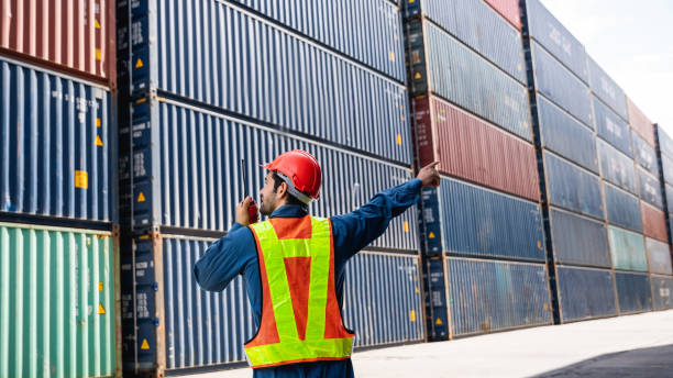 istock Engineer or foreman standing on the container yard warehouse talk on a portable radio and check inventory or task details., Logistics concept inside the shipping, import, and export industries. 1455899996