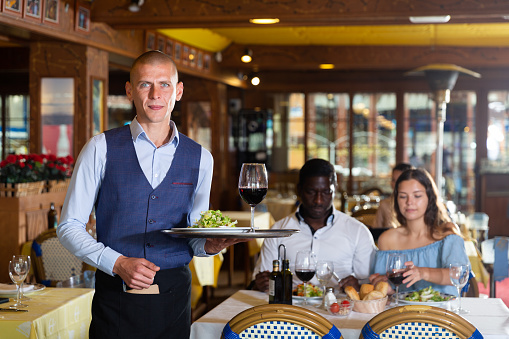 Professional young smiling waiter holding serving tray for restaurant guests