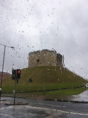 View of Clifford's Tower in York, UK with road and traffic light from wet glass window with rain drop during storm with cloudy sky background. No people.