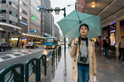 smiling asian Japanese girl tourist holding umbrella and looking into distance at the cityscape while taking stroll on the pedestrian walk on shijo dori in downtown Kyoto japan in the evening