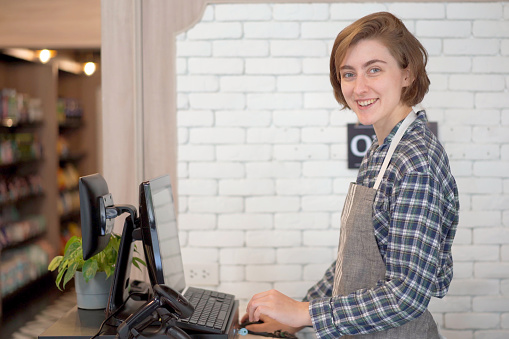 Caucasian white female shopkeeper standing behind a cashier counter and smiling to camera. Happy female shopkeeper or coffee shop staff looking and smiling at camera.