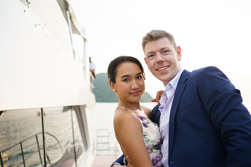 Happy Asian and caucasian couple dating on a yacht together. Lovely couple in love staying on yacht corridor and watching sunset together.