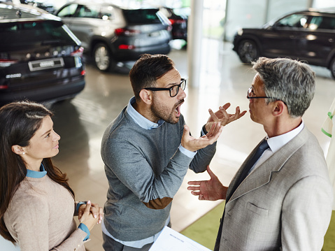 Displeased man and his wife arguing with a salesman in a car showroom.