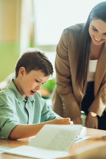 Elementary schoolboy looking happy after finishing his exam paper while a female teacher sitting by and smiling. Elementary school students in classroom with teacher.