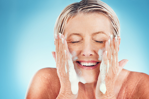 Face, cleaning and senior woman washing, skin and water splash for wellness, beauty and skincare in a studio. Beauty splash, facial and elderly wellness model happy, smile and relax with soap product