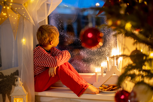 Toddler child, cute blond boy, sitting on the window in pajama, looking out for Satna Claus,, eating cookies,  Christmas lights around him