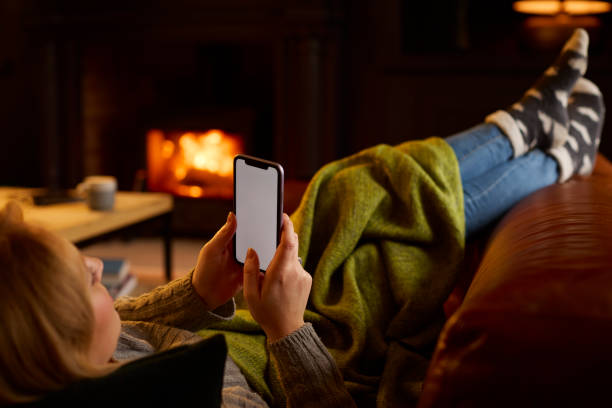 istock Woman At Home Lying On Sofa In Lounge With Cosy Fire Using Blank Screen Mobile Phone 1455926506