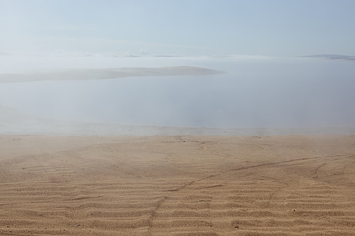 Lonely lifeless lake shore in early morning with soft haze above water and yellow sand coast with cape in fog. Mysterious and unearthly landscape.