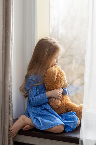 Sad little girl sits on the windowsill with her teddy bear and looks out the window.